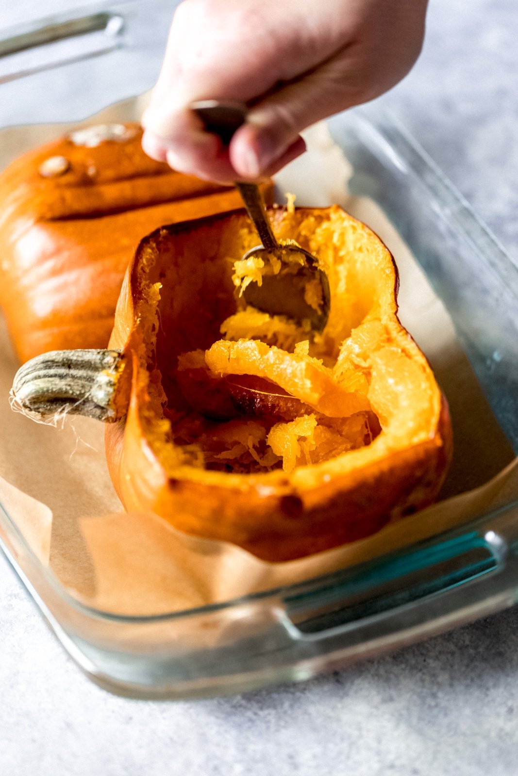 hand scooping out the inside of half of a cooked pumpkin in a glass baking dish