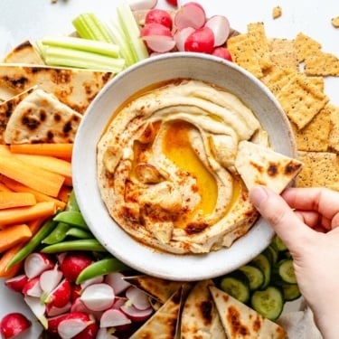 dipping pita into a bowl of roasted garlic hummus surrounded by veggies